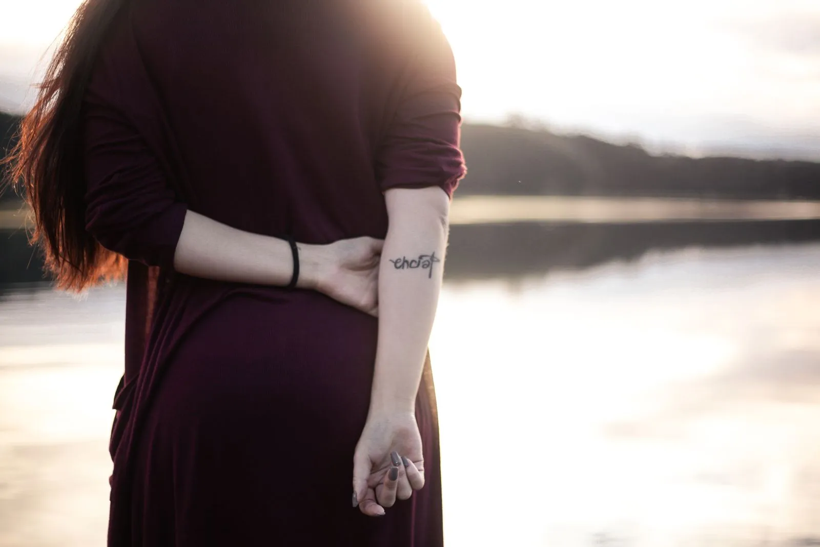 Back view of a woman in a brown dress standing by a calm lake at sunset.